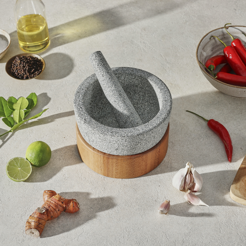 empty granite mortar and pestle on wooden base surrounded by garlic, chilli, lime and galangal